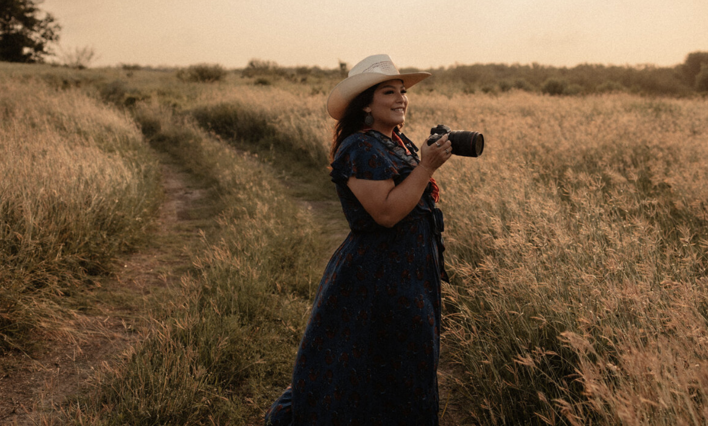 Wedding photographer holding a camera and taking photos in the middle of a field during a wedding or engagement session, capturing natural light and scenic surroundings.