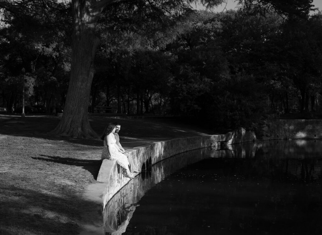 Black and white portrait of an engaged couple sitting beside the pond during a peaceful sunset engagement session at the San Antonio Botanical Garden.