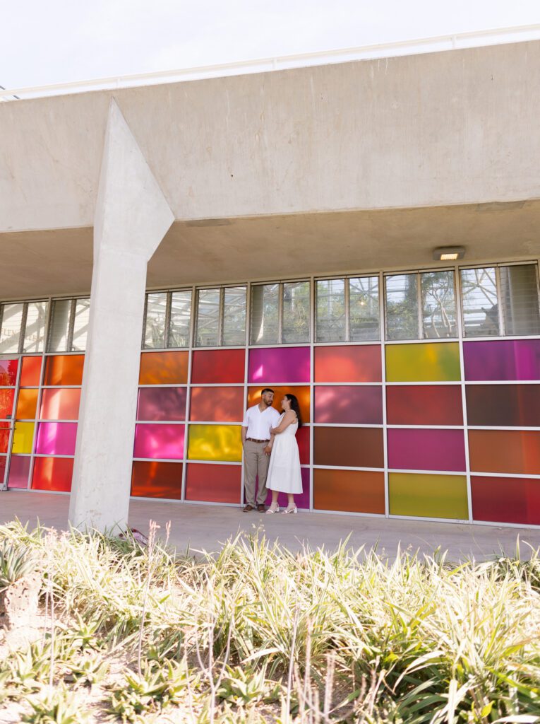 Wide portrait of an engaged couple standing beside the colorful glass conservatory at the San Antonio Botanical Garden.