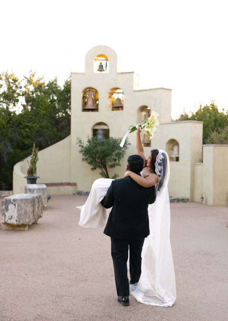 Groom carrying his bride beneath the iconic bell wall at Chapel Dulcinea in Austin, Texas.